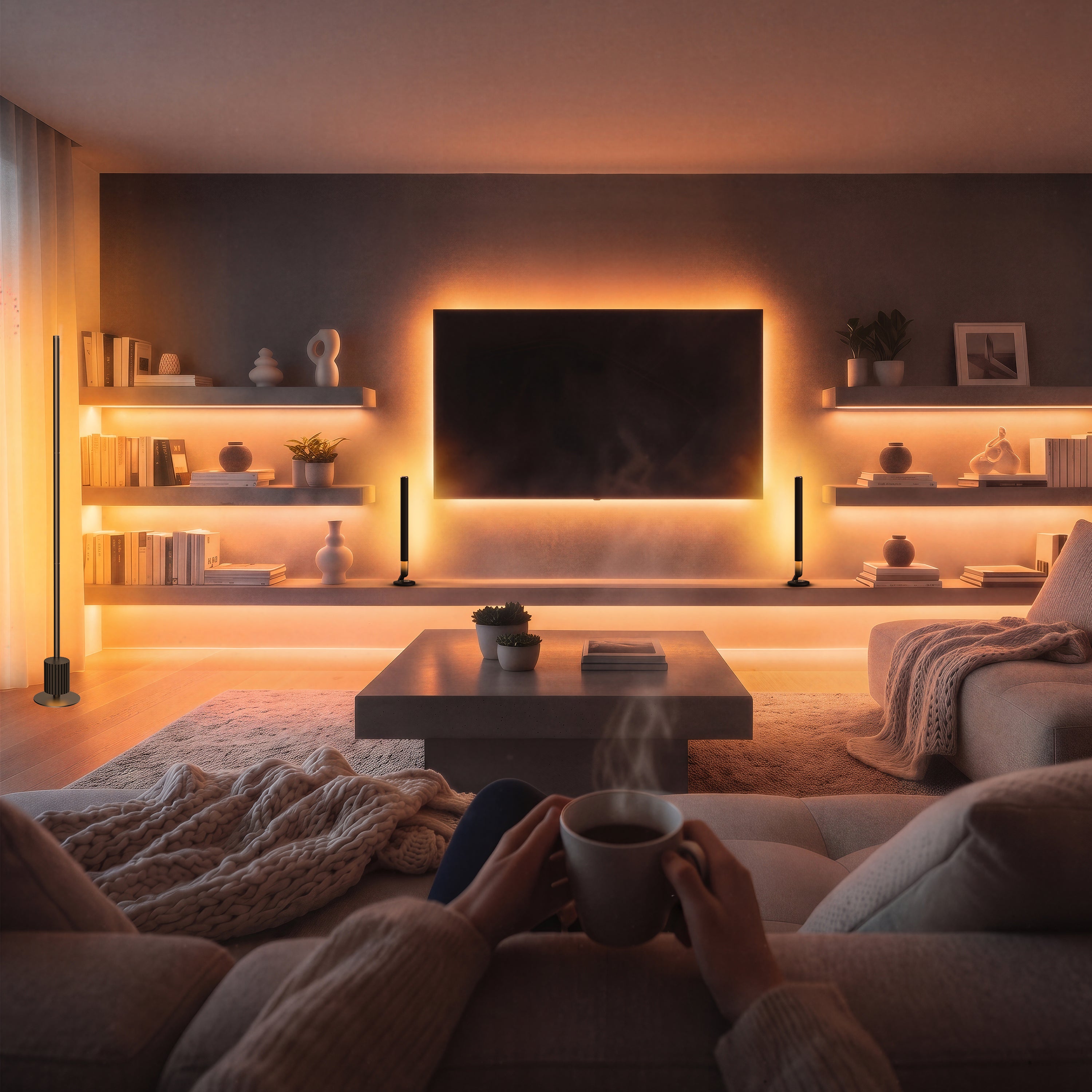 A cozy living room scene viewed from a couch, with a person holding a steaming mug in their hands. The room features a dark gray wall with built-in shelves, warm under-shelf lighting, a black wall-mounted TV, and decorative objects.
