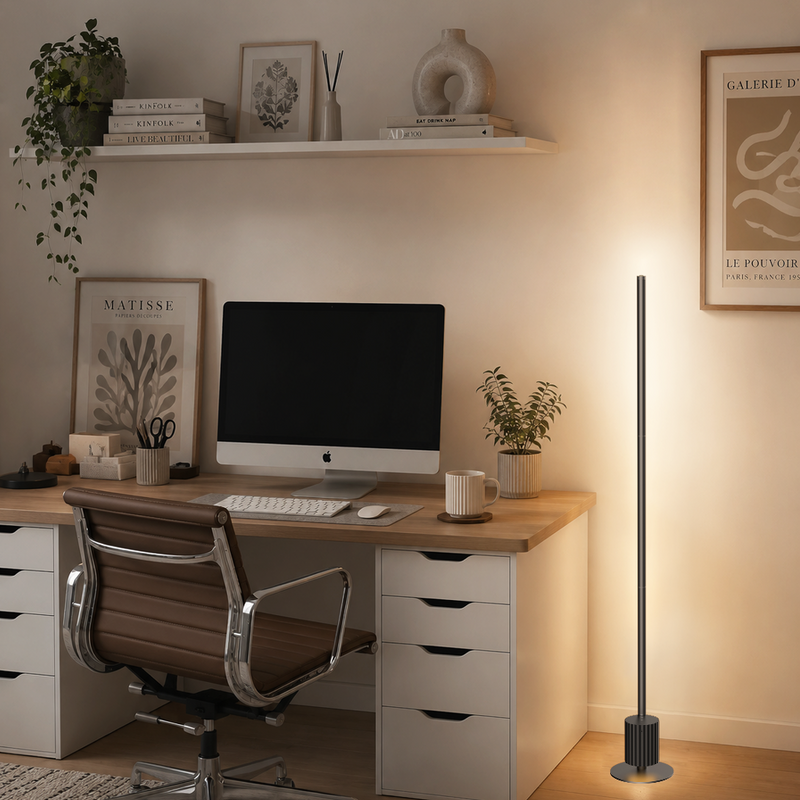 A modern minimalist home office scene featuring a slim silver floor lamp on the right, casting a warm glow against a beige wall. The room includes a light wood desk with white drawers, a brown leather office chair, a computer monitor, and various decorative items on a floating shelf.
