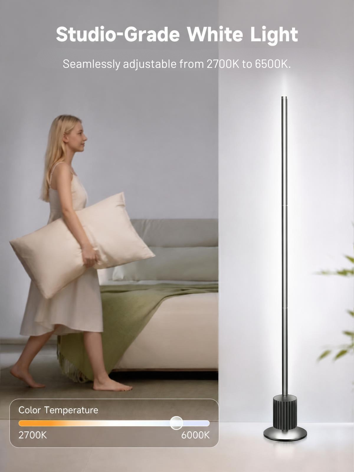 A tall slim floor lamp with a metallic silver base and pole stands on the right against a light wall, while a woman in a cream dress carrying a pillow walks past a bed on the left. Text at the top reads 'Studio-Grade White Light' and 'Seamlessly adjustable from 2700K to 6500K.