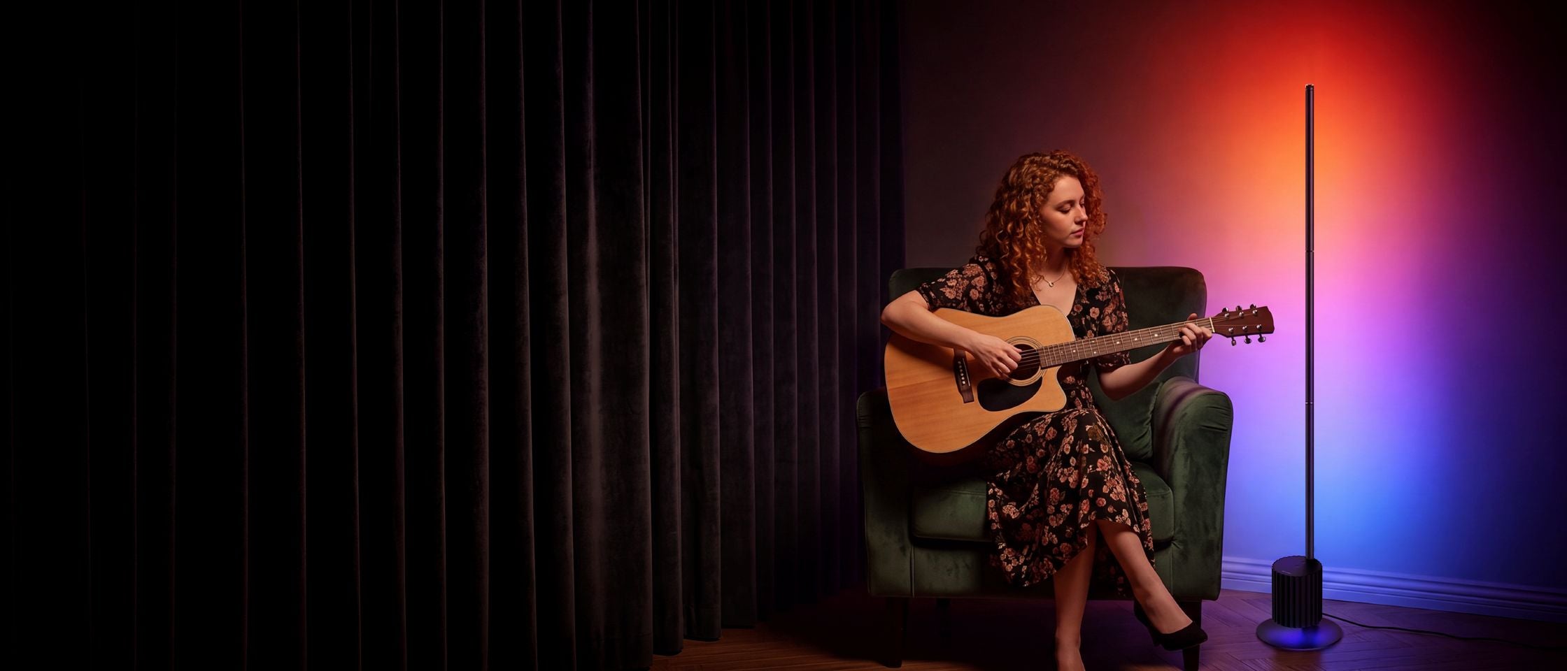 A woman with curly hair sits in a green armchair, playing an acoustic guitar in a dimly lit room with a black curtain on the left and a floor lamp casting red, orange, and blue light on the right.